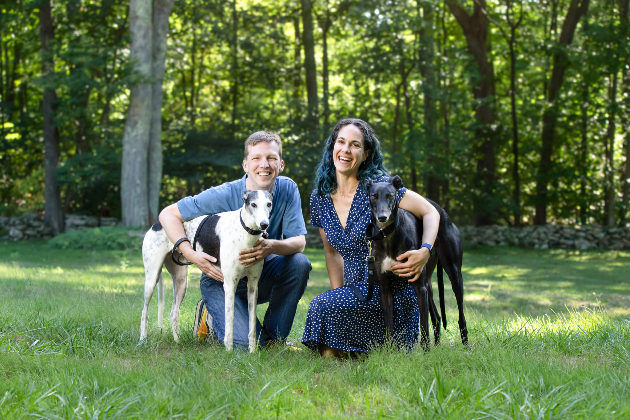 A married couple each hug their two large dogs while kneeling in a field in front of a tree line